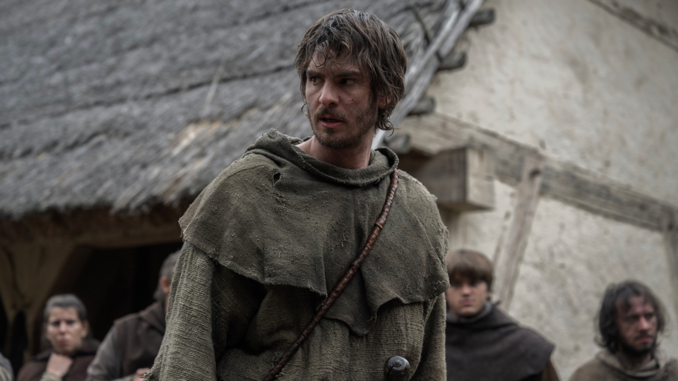 Andrew Garfield, com um capuz de tecido rústico e barba por fazer, olha para o lado numa aldeia medieval em "A Revolta"